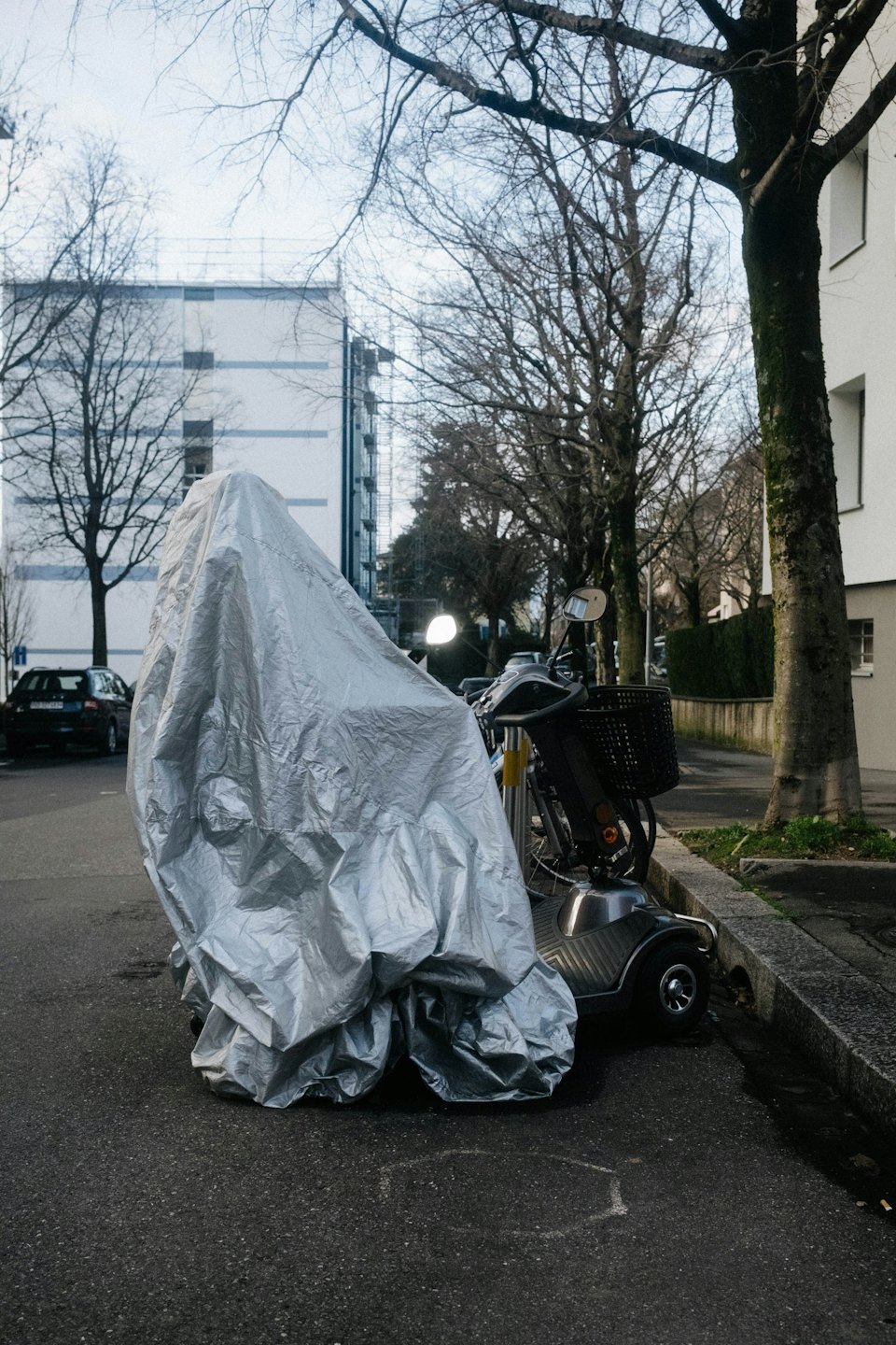 Silberner Elektroroller unter einer Plane auf Asphalt.